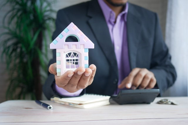 A man in a business suit, holding a small house in one hand and performing calculations on a calculator with his other hand