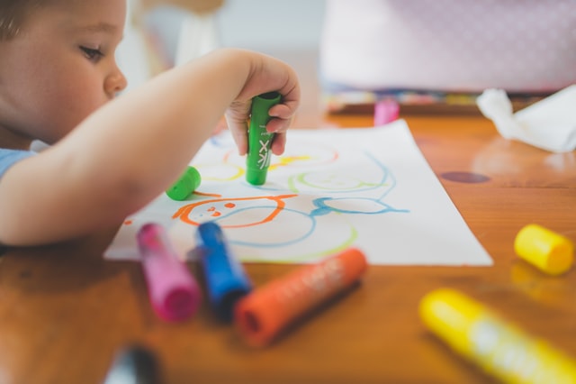 A boy sitting at a desk and drawing on white paper with crayons