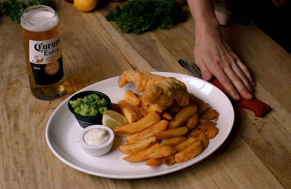 A glass of beer beside a plate of fish and chips.