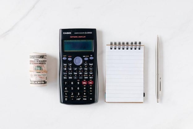 A stack of money on the left, a calculator in the middle, and a notebook and a pen on the right