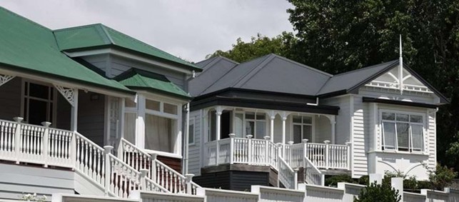 Row of white wooden houses with wraparound porches and decorative trim, varying green/gray roofs in a sunny neighborhood