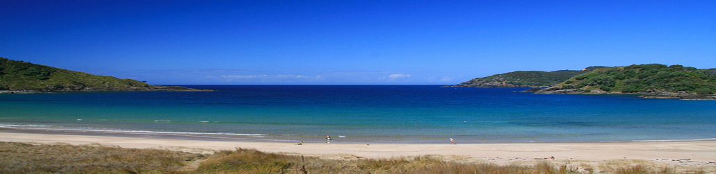 Sunny coastal scene with a sandy beach, turquoise sea, and green hills in the background under a blue sky.